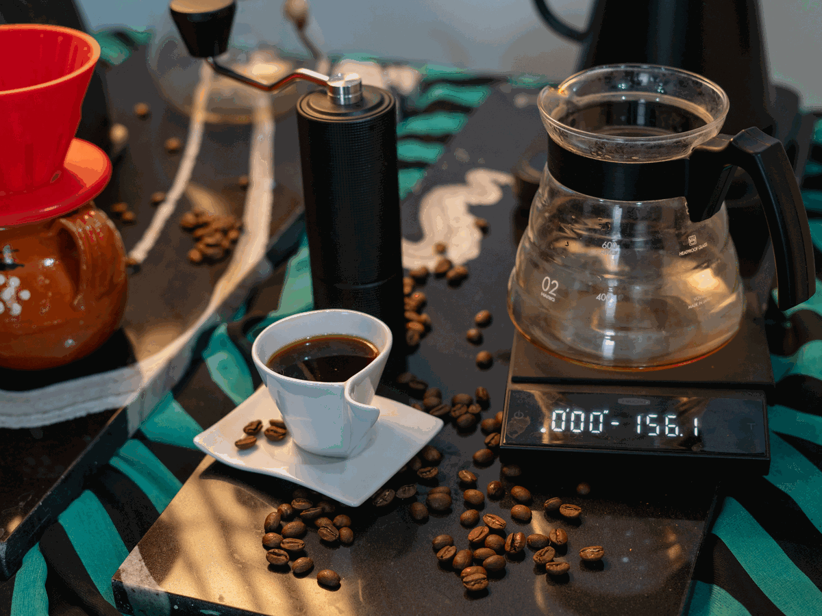 深色桌面上擺著黑色手搖磨豆機、紅色濾杯、玻璃咖啡壺和電子秤，前面一杯黑咖啡，旁邊散著咖啡豆
